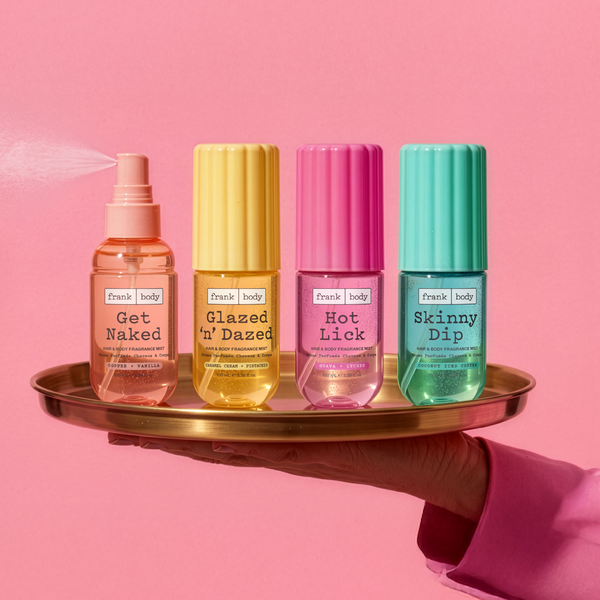 The four frank body hair and body mists — Get Naked, Glazed 'n' Dazed, Hot Lick and Skinny Dip — on a gold tray, one mid-spray.