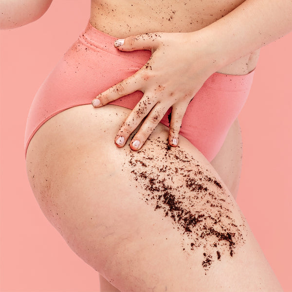 A close-up of a person wearing pink underwear, with dark brown scrub or powder applied to the upper thigh and hand, against a solid pink background.