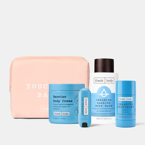 A Frank Body travel care set featuring a light pink zippered pouch, a blue Barrier Body Creme jar, a white and blue Barrier Body Wash bottle, a blue deodorant stick, and a blue body spray, arranged on a light gray background.