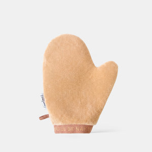 A beige plush bath mitt with a brown elastic wrist strap and a small fabric tag, shown on a light gray background.