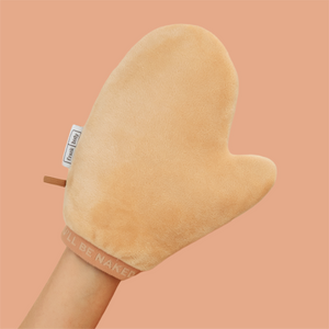 A plush peach-colored silicone or fabric hand mitt with a handle, worn on a person's hand against a solid peach background. The mitt features a small white tag reading 'frank body' and a pinkish-beige cuff with the text 'I'LL BE NAKED'.