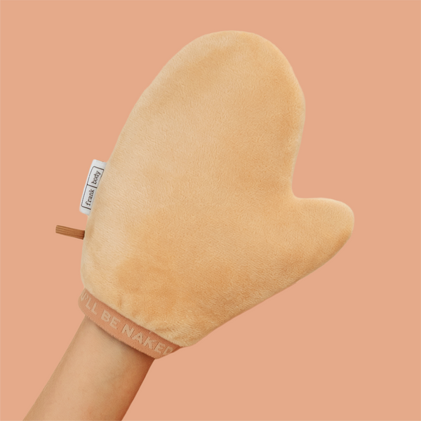 A plush peach-colored silicone or fabric hand mitt with a handle, worn on a person's hand against a solid peach background. The mitt features a small white tag reading 'frank body' and a pinkish-beige cuff with the text 'I'LL BE NAKED'.