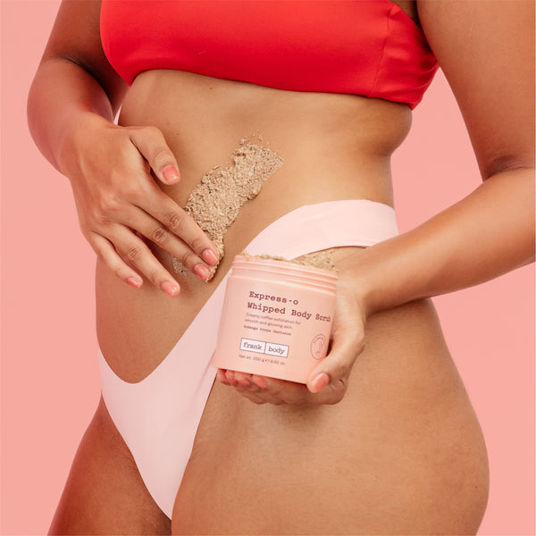 A woman in a red bikini top and pale pink bikini bottom applies a beige whipped body scrub from a pink tub labeled 'Express-o Whipped Body Scrub' against a solid pink background.