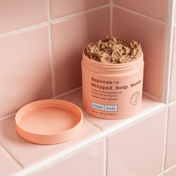 A peach-colored jar of Express-o Whipped Body Scrub sits on a pink tiled surface with its lid placed beside it. The scrub itself is a thick, textured brown.
