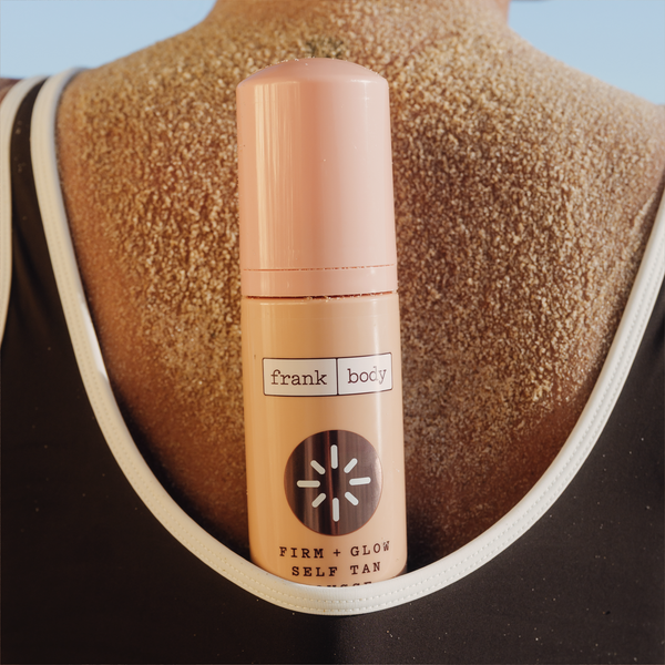 A bottle of frank body Firm + Glow Self Tan Mousse resting against a tanned shoulder.