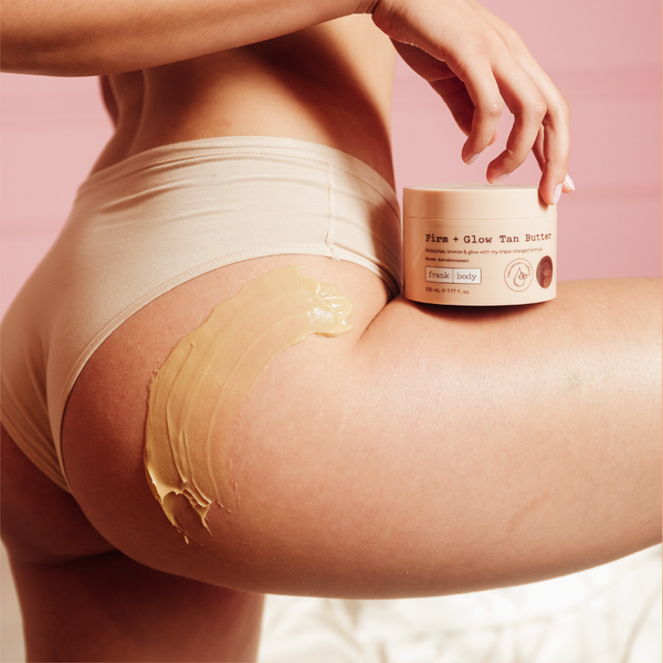 A close-up lifestyle shot of a model wearing beige underwear with a thick, glossy tan body butter applied to her upper thigh. She is holding a jar of 'Firm + Glow Tan Butter' against a solid pink background.