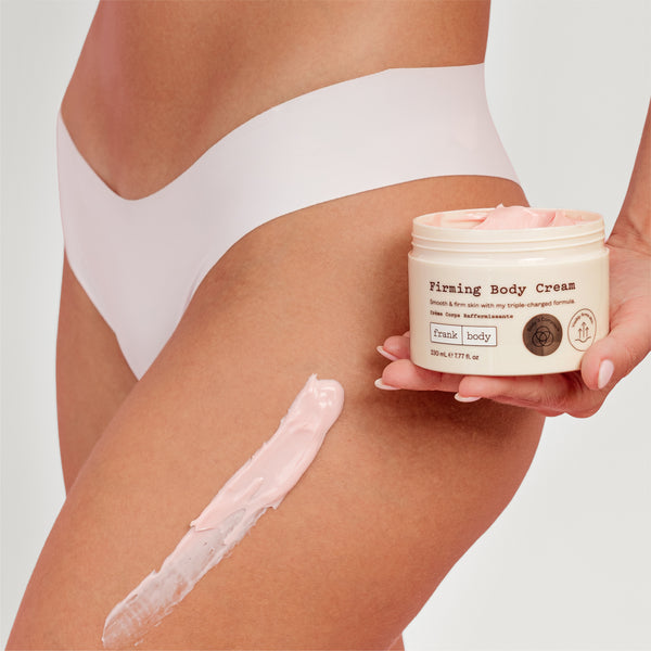 A person wearing white underwear holds a jar of Firming Body Cream next to a smear of pale pink cream on their thigh against a light gray background.