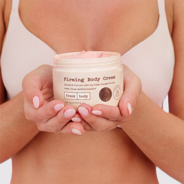 A close-up of a person holding a jar of Firming Body Cream. The person is wearing a light pink bikini top, and the cream-colored jar is held in their hands against a clean white background.
