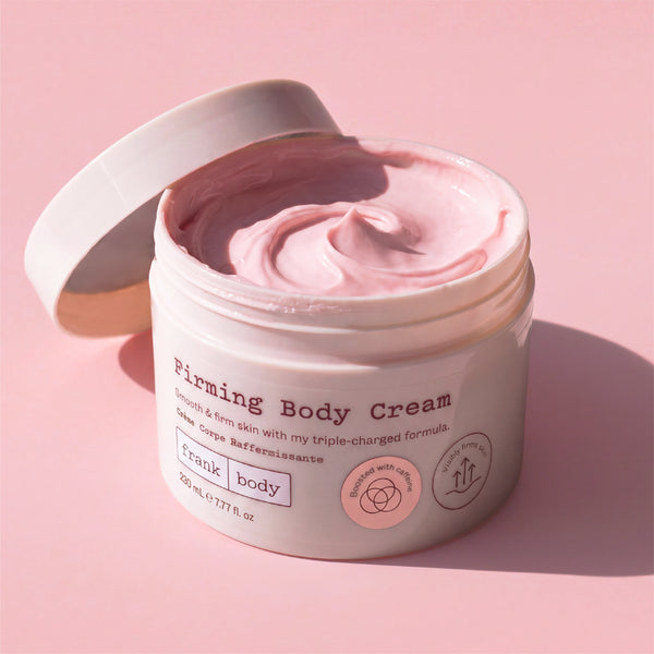 An open white jar of Frank Body Firming Body Cream, showing the pink cream inside, set against a solid pink background with a soft shadow.