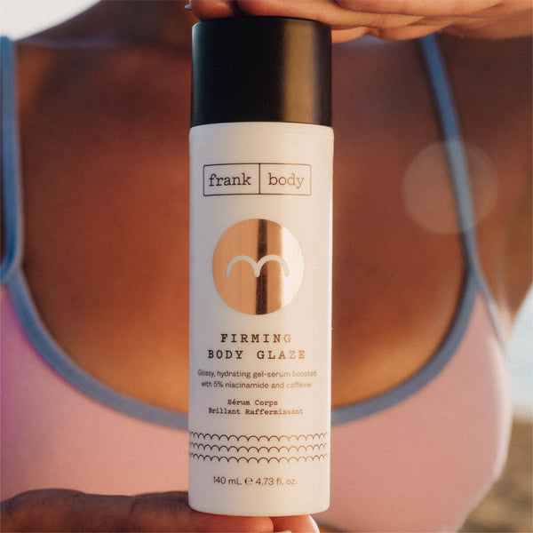 A white bottle of Frank Body Firming Body Glaze with a black cap, held in front of a blurred person wearing a pink top.