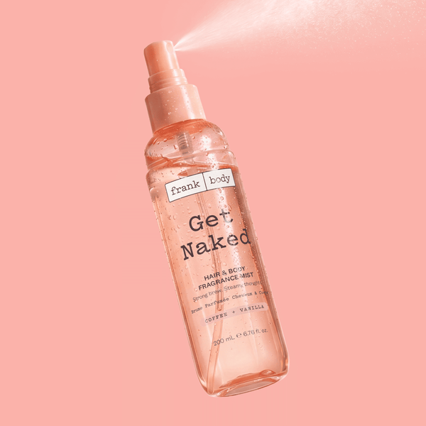 A bottle of frank body Get Naked Hair & Body Fragrance Mist mid-spray on a pink background.