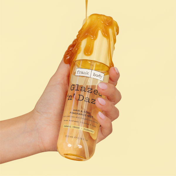 A hand holding a bottle of frank body Glazed 'n' Dazed Hair & Body Fragrance Mist with caramel dripping down it.