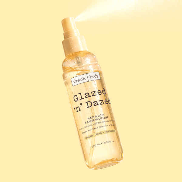 A bottle of frank body Glazed 'n' Dazed Hair & Body Fragrance Mist mid-spray on a yellow background.