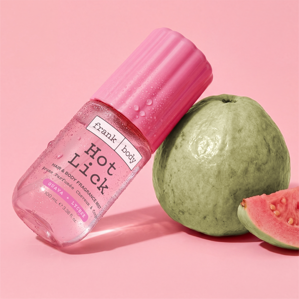 A pink bottle of frank body On-the-Go Hot Lick Hair & Body Fragrance Mist beside a whole and halved guava.