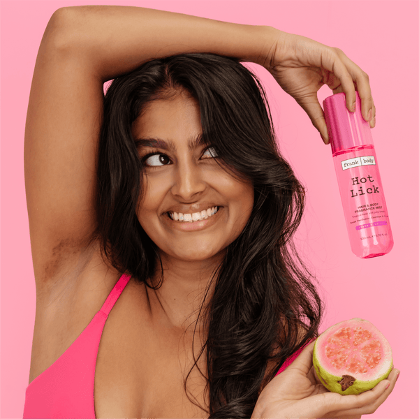 A smiling woman in a pink bra holding a bottle of frank body Hot Lick Hair & Body Fragrance Mist and half a guava.