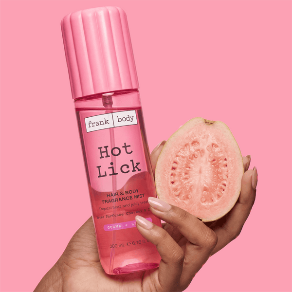 A hand holding a bottle of frank body Hot Lick Hair & Body Fragrance Mist beside half a guava.