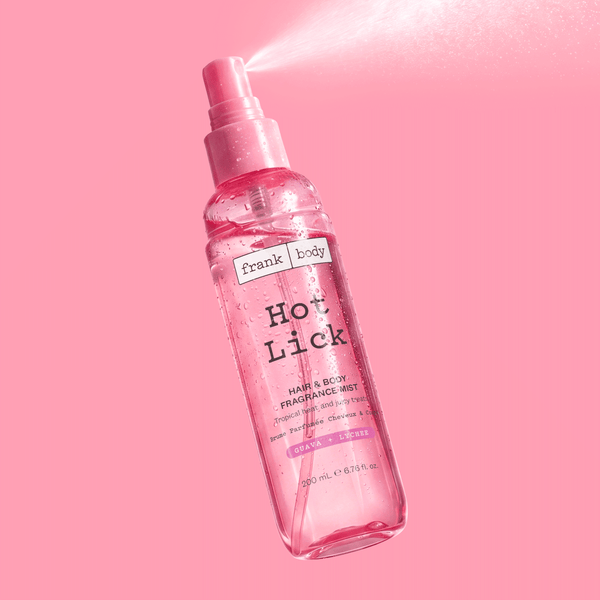 A bottle of frank body Hot Lick Hair & Body Fragrance Mist mid-spray on a pink background.