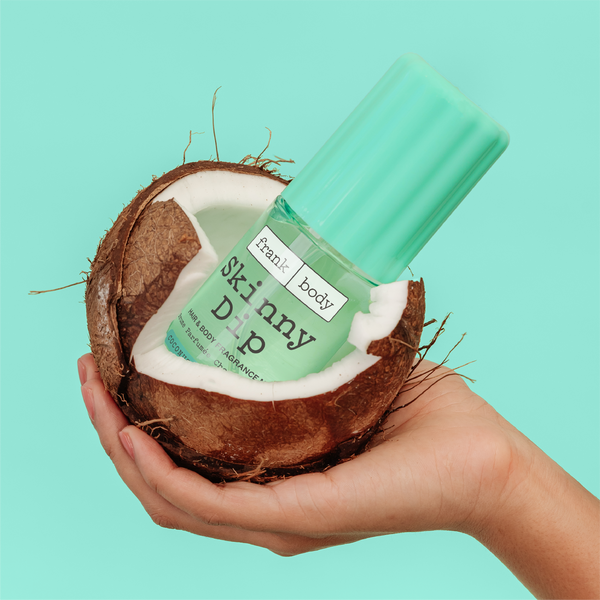 A hand holding a bottle of frank body Skinny Dip hair and body mist inside a cracked coconut.