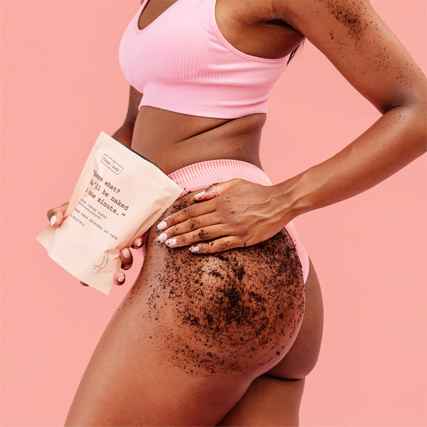 A woman in a pink ribbed sports bra and matching shorts holds a beige paper pouch labeled "Original Coffee Scrub" while applying a dark brown scrub to her upper thigh. The background is a solid pastel pink.