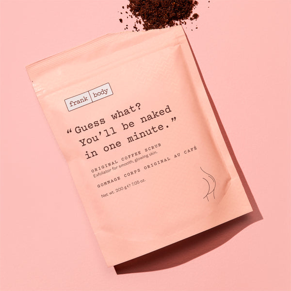 A pink stand-up pouch of Frank | Body Original Coffee Scrub with styled on a matching pink background with coffee grounds scattered at the top.