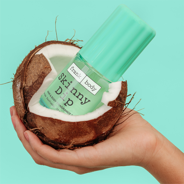 A bottle of frank body On-the-Go Skinny Dip Hair & Body Fragrance Mist resting inside a cracked coconut.
