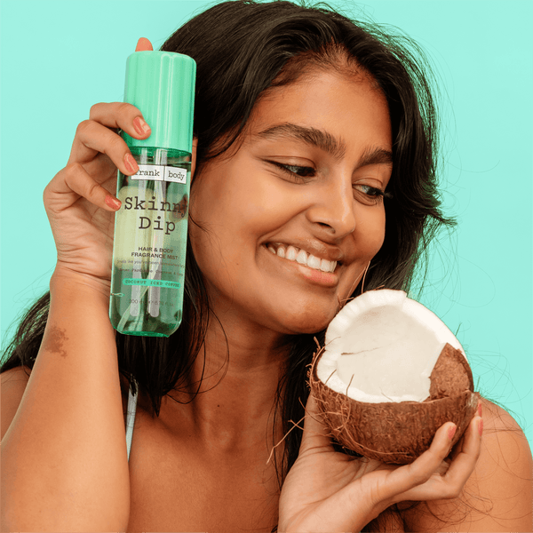 A smiling woman holding a bottle of frank body Skinny Dip Hair & Body Fragrance Mist beside half a coconut.