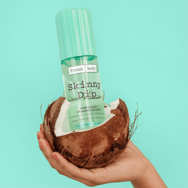 A bottle of frank body Skinny Dip Hair & Body Fragrance Mist resting inside a cracked coconut.