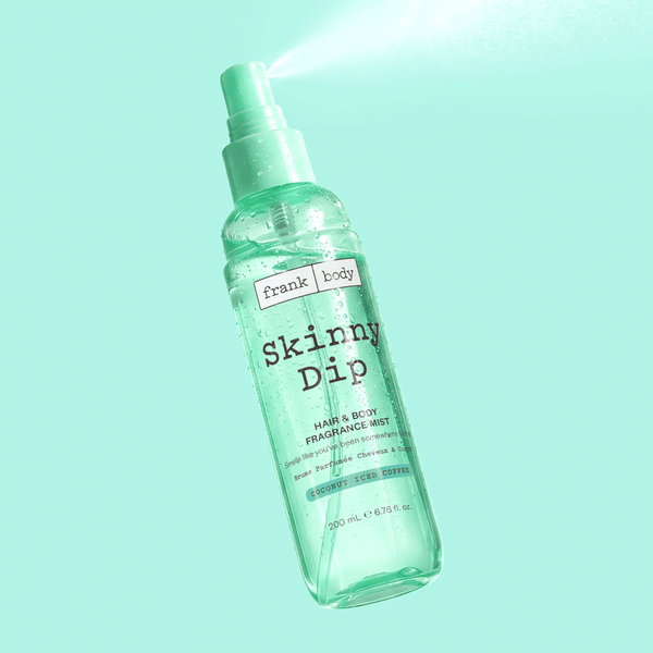 A bottle of frank body Skinny Dip Hair & Body Fragrance Mist mid-spray on a mint green background.