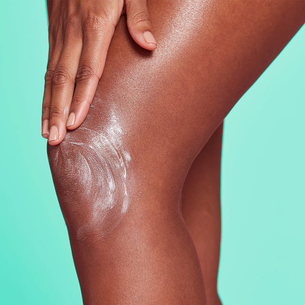 Close-up of a person's knee and hand applying or smoothing a white cream or lotion onto the skin against a bright aqua background.