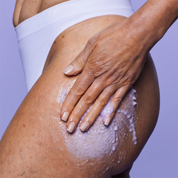 A close-up of a person's hip and upper thigh with a white cream or exfoliating scrub being rubbed in by a hand. The person is wearing white underwear against a solid light purple background.