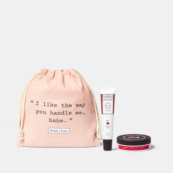 The frank body Tis The Cherry Season Kit: Cherry Bomb Lip Tint and Cherry Bomb Lip Scrub in a pink drawstring bag.