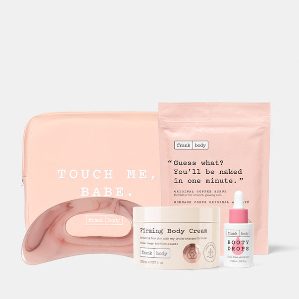 A Frank Body self-care set featuring a blush pink pouch with 'TOUCH ME, BABE.' text, a matching pink curved tray, a cream jar labeled 'Firming Body Cream,' a small white bottle labeled 'BOOTY DROPS,' and a pink stand-up pouch with a quote, arranged on a light gray background.