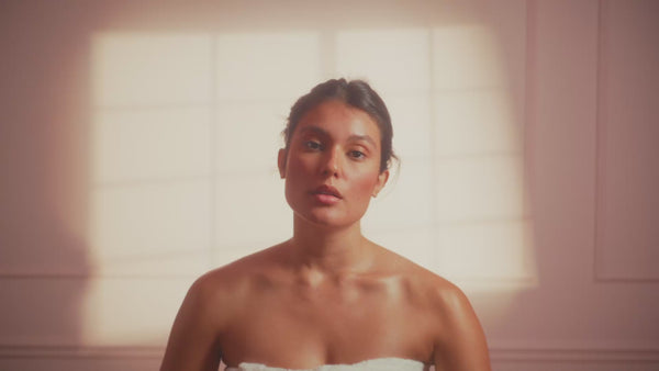 frank body Firm + Glow Self Tan Mousse video: a woman wrapped in a towel in a sunlit bathroom applying the self tan mousse.