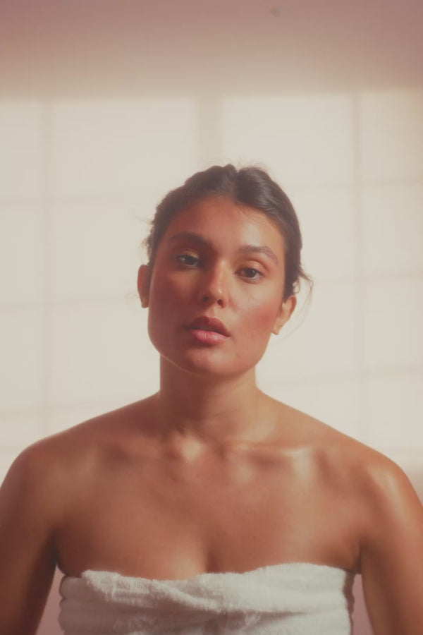 frank body Firm + Glow Self Tan Mousse video: a woman wrapped in a towel in a sunlit bathroom applying the self tan mousse.